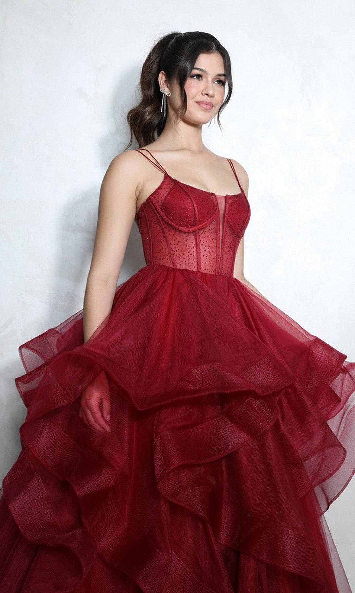 A woman poses against a light background, wearing the Eureka 9950 long prom dress in burgundy, featuring a layered ballgown skirt and a fitted, sheer beaded corset bodice with thin straps.